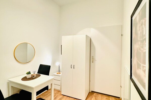 A small room with a white desk, a chair, a round mirror, a wardrobe, and a framed picture on the wall.