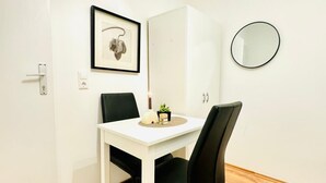 A white dining table with two black chairs, a framed picture on the wall, a round mirror, and a small potted plant on the table.