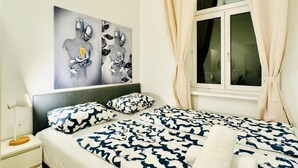A bedroom with a bed, two pillows, a nightstand, a lamp, and a window with curtains.