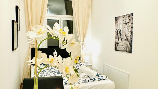 A hotel room with a bed, a vase of white flowers, a black chair, and a framed picture on the wall. - Modern 2-Person Room, Close to Atzgersdorf (Vienna)