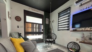 Una sala de estar moderna con un sofá gris, un cojín amarillo, una silla colgante, un ventanal con persianas y un televisor de pared.