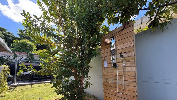 Ducha exterior de madera con manguera y cabezal de ducha manual, rodeada por un jardín cubierto de césped y un árbol.