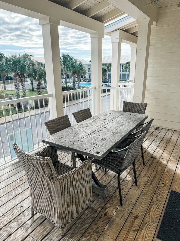 A covered outdoor dining area with a wooden table and wicker chairs, overlooking a pool and buildings.
