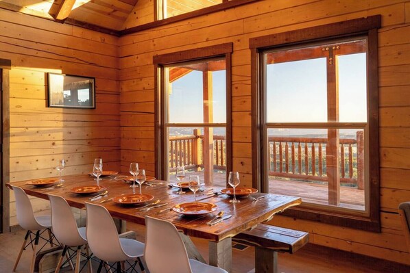Een houten eetkamer met een grote tafel gedekt voor zes personen, grote ramen en uitzicht op een houten terras.
