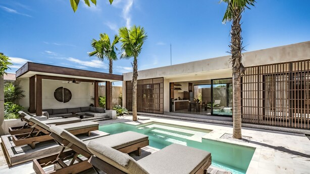 Uma área moderna com piscina ao ar livre, espreguiçadeiras, palmeiras e um edifício contemporâneo com amplas janelas.