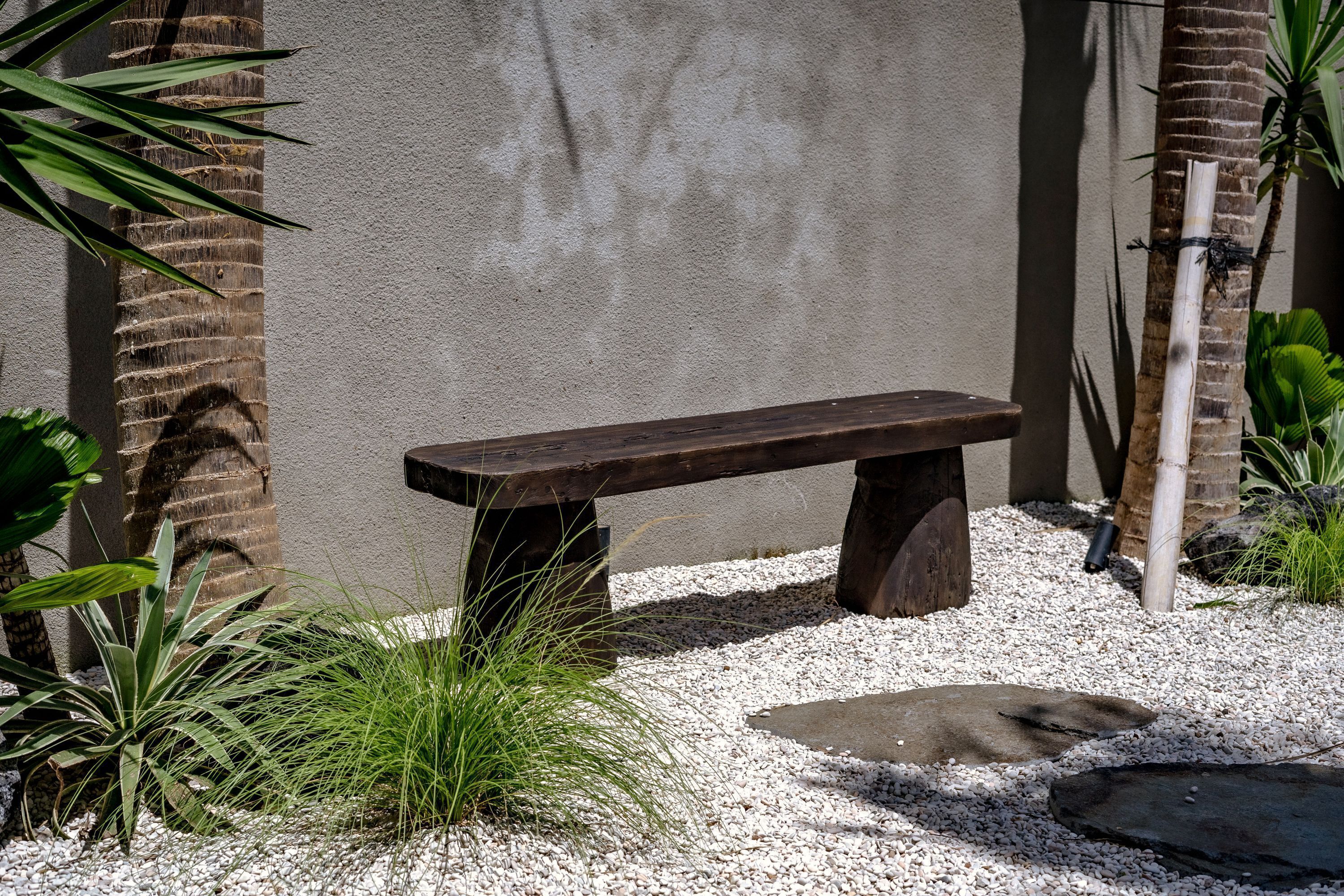 A wooden bench in a garden area with gravel, surrounded by plants and a concrete wall.