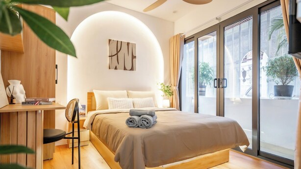 A modern bedroom with a bed, a desk, and a chair. There is a large window with curtains, and a plant is visible outside.