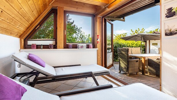 Une véranda avec un plafond en pente, un banc de fenêtre agrémenté de coussins et un espace patio avec des meubles d’extérieur.