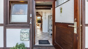 Une porte en bois avec un paillasson, une fenêtre ornée d’un cadre décoratif et une vue sur une pièce dont le sol est recouvert de carreaux.