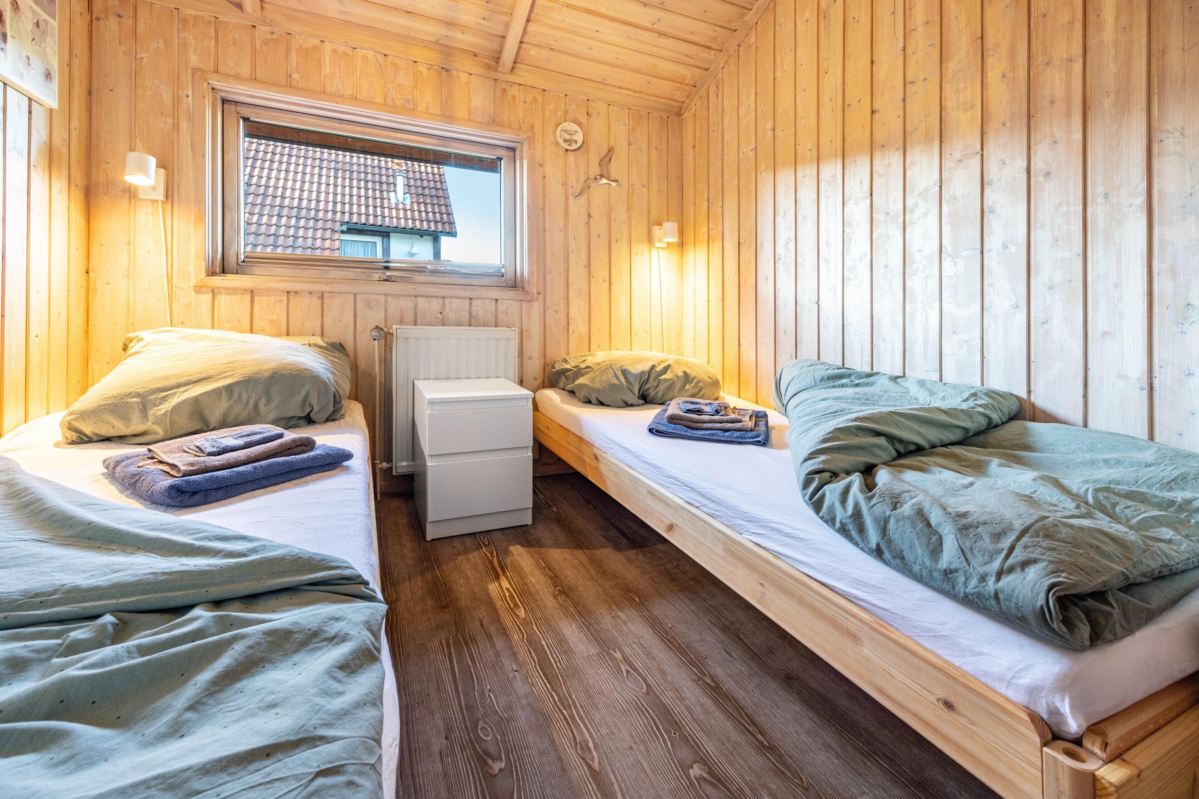A wooden cabin with two single beds, a small white dresser, and a window with a view of a tiled roof.