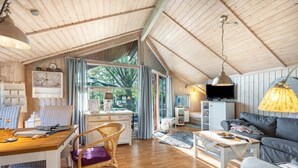 Een ruime kamer met houten balkenplafond, een eethoek met tafel en stoelen, een bank, een televisie en een groot raam met gordijnen.