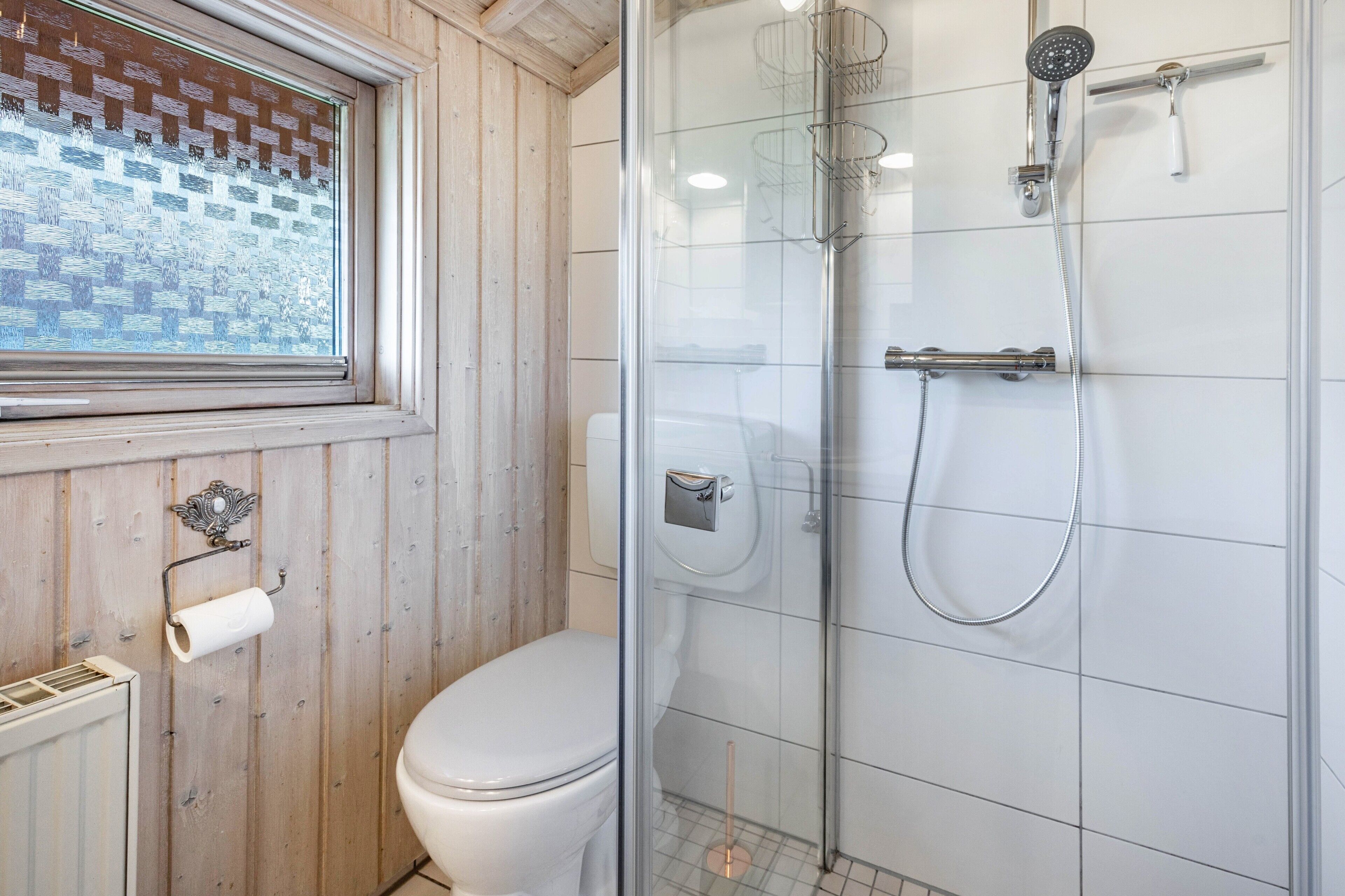 Un baño con inodoro, ducha con cerramiento de vidrio y ventana con diseño de ladrillo.
