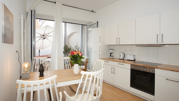 A modern kitchen with white cabinetry, a wooden countertop, and a built-in oven. There is a dining area with white chairs and a table, and a window with a view of the outdoors. - Apartment 'Fewo Romeo und Julia' with Private Terrace and Wi-Fi (Bamberg)