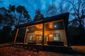 Une cabane moderne en bois, à deux étages, dotée d’un porche couvert, de grandes fenêtres et d’un lustre central.