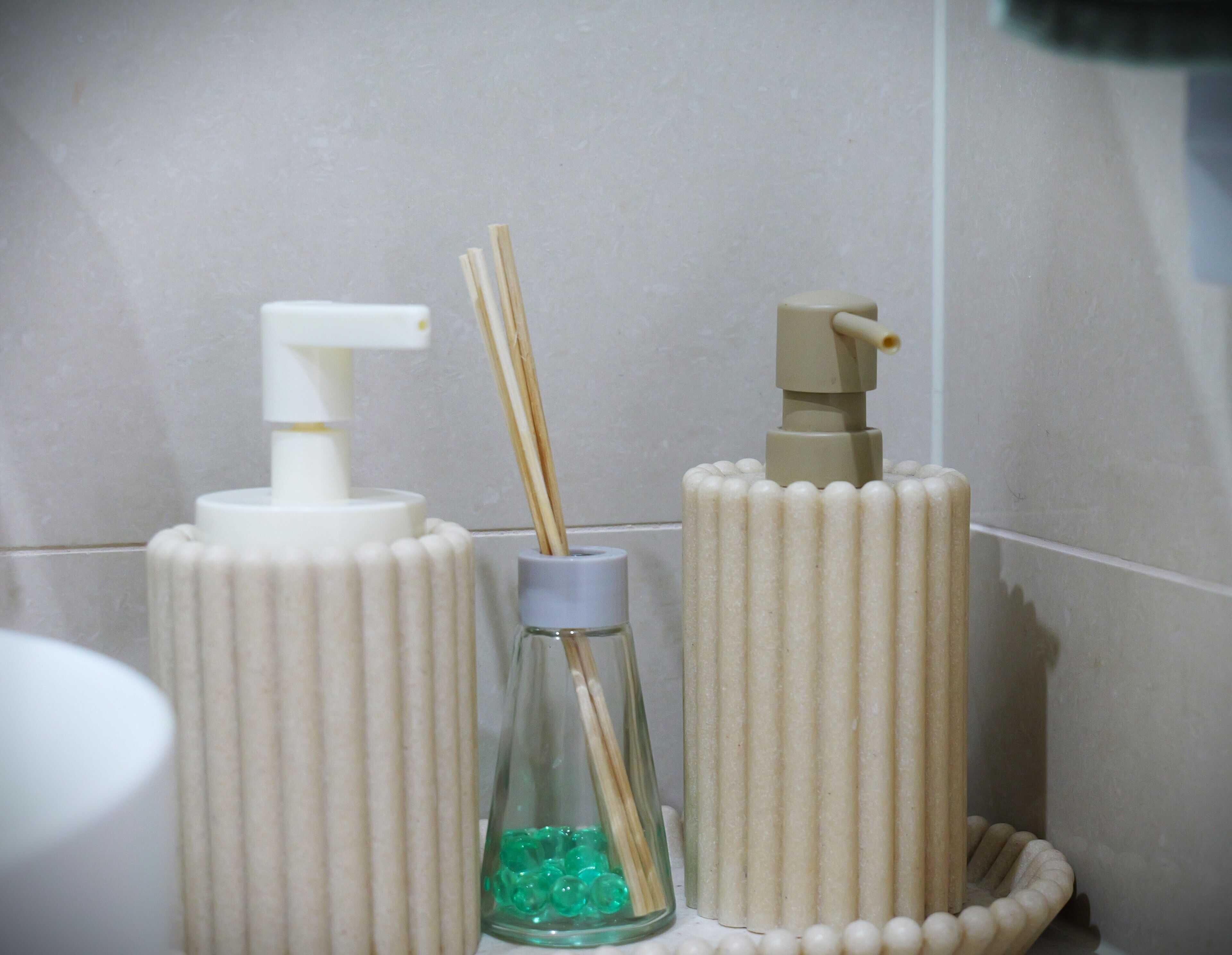 Une salle de bain équipée de deux distributeurs cylindriques, l’un avec une pompe et l’autre avec un bouton-poussoir, ainsi que d’un flacon en verre contenant un diffuseur à tiges.