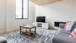 En moderne stue med et stort vindue, et fladskærms-tv på et hvidt skab, en grå sofa med mønstrede puder og et træbord.