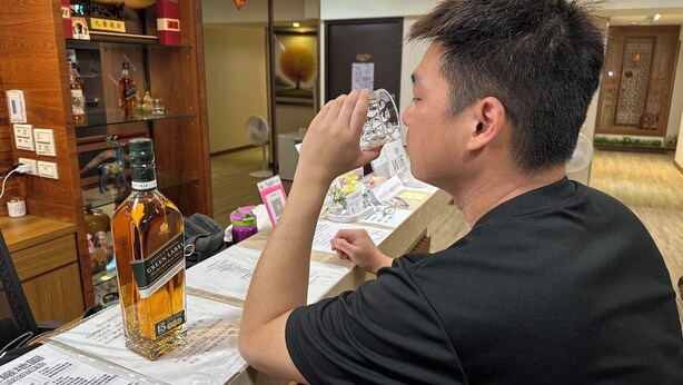 Un hombre sentado a una mesa con una botella de whisky Green Label, un menú y varios objetos sobre ella.