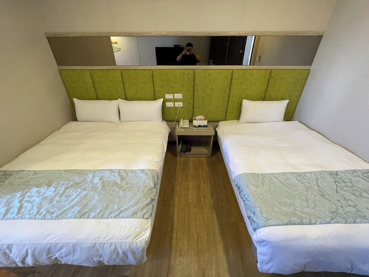 Habitació d'hotel amb dos llits, un capçalera verd, una tauleta de nit amb un telèfon i mocadors, i un mirall que reflecteix una persona.