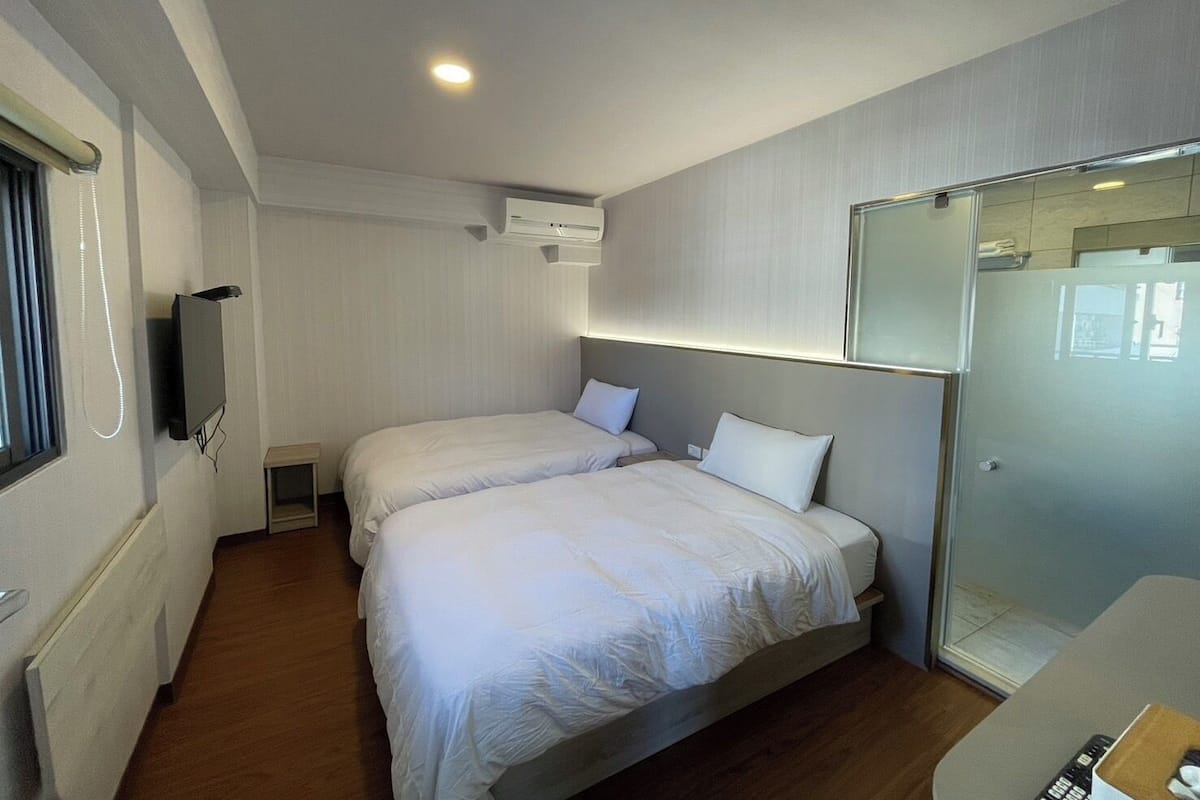 Een hotelkamer met twee bedden, een televisie, een airconditioning en een douchecabine van glas.