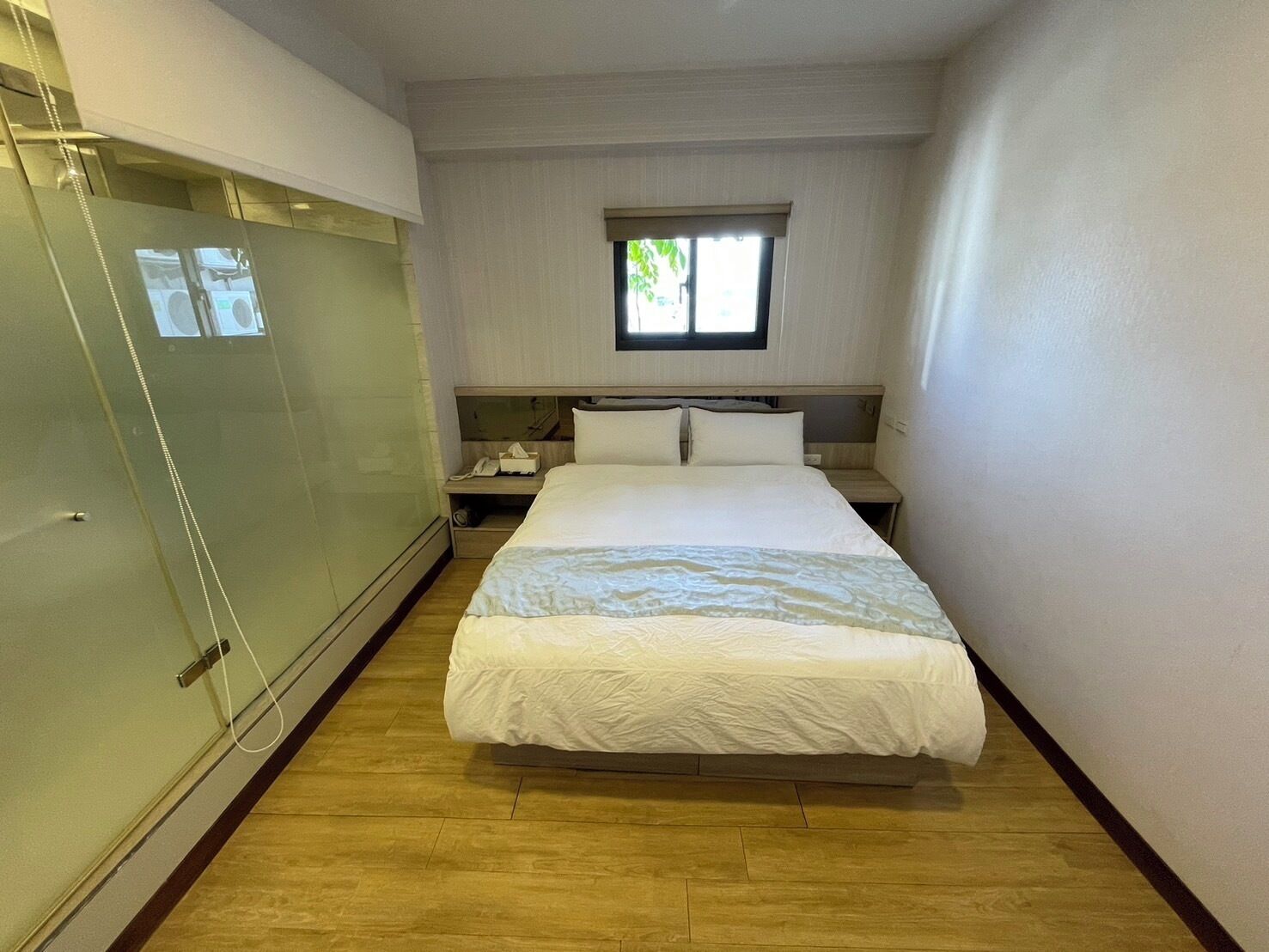 A hotel room with a bed, a nightstand, a window, and a glass door.