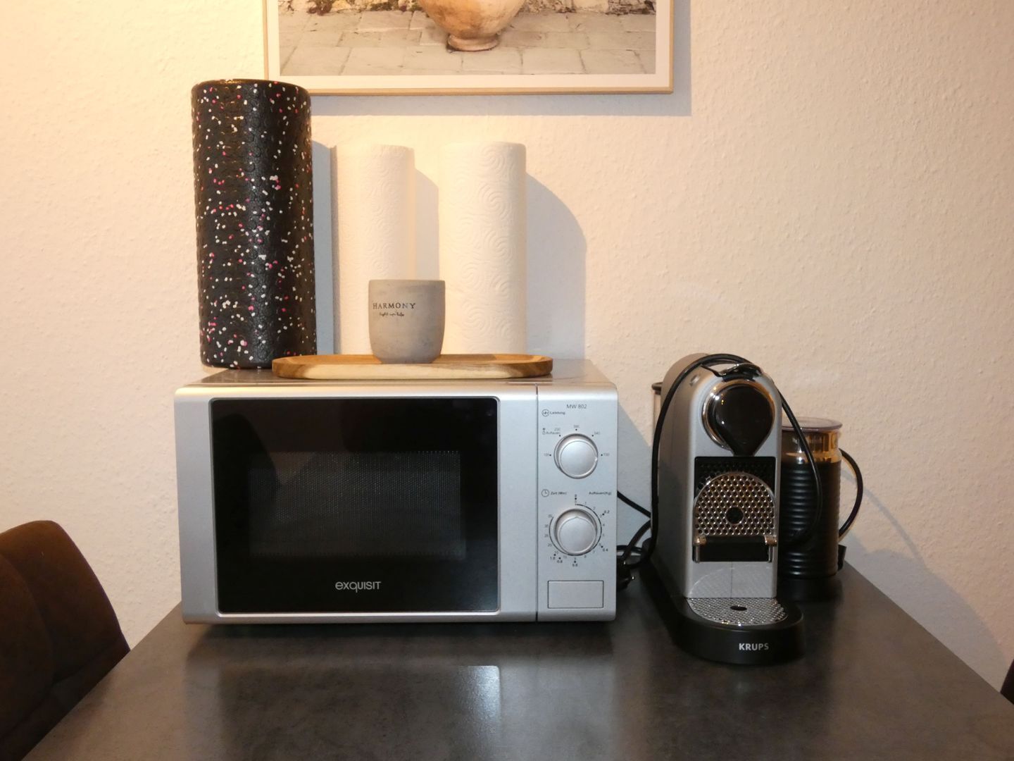 Eine Mikrowelle mit Holztablett und einer kleinen Tasse darauf, ein Wasserkocher und ein gerahmtes Bild an der Wand.