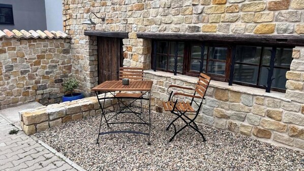 Een stenen gebouw met een patio, voorzien van een houten tafel en stoelen, een kleine potplant en een raam met donkere kaders.