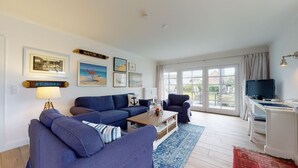A living room with a blue sofa set, a coffee table, and a TV on a stand. There are framed pictures on the wall, a lamp, and a sign that reads "JESSIE". - Modern apartment with terrace and sea view. - At the dune (Hörnum (Sylt))
