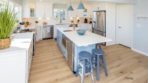 A modern kitchen with white cabinetry, a central island with stools, stainless steel appliances, and pendant lighting. - Ebb's-Oceanfront, Direct Beach Access, Pool, Sleeps 16! (Virginia Beach)