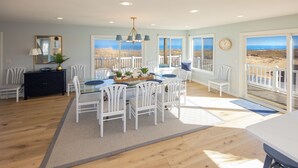 A dining area with a large table, white chairs, and a view of the beach through large windows. - Ebb's-Oceanfront, Direct Beach Access, Pool, Sleeps 16! (Virginia Beach)