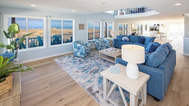 A spacious living room with a large window overlooking the beach, a blue sofa, and a white coffee table. - Ebb's-Oceanfront, Direct Beach Access, Pool, Sleeps 16! (Virginia Beach)