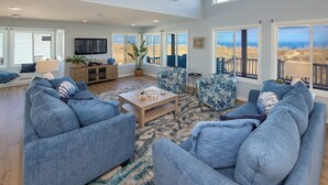 A spacious living room with a large blue sofa set, a wooden coffee table, and a view of the beach through large windows. - Ebb's-Oceanfront, Direct Beach Access, Pool, Sleeps 16! (Virginia Beach)