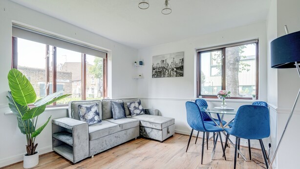 Ein modernes Wohnzimmer mit einem grauen Ecksofa, einem Esstisch mit blauen Stühlen, großen Fenstern und einer Topfpflanze.