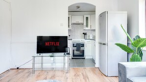 Ein modernes Wohnzimmer mit einem Flachbildfernseher, einem Glastisch und einer Couch. Im Hintergrund ist eine Küche mit weißen Schränken und einem weißen Kühlschrank zu sehen.