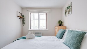 Ein Schlafzimmer mit einem Bett, einem Fenster, einem an der Wand befestigten Regal mit einer Pflanze, einem Nachttisch ebenfalls mit einer Pflanze und einem Kunstwerk an der Wand.