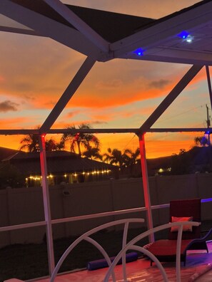 A covered outdoor seating area with a view of a sunset over palm trees.