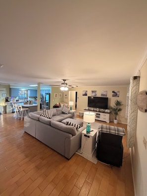Un salon avec un canapé gris, un téléviseur, un ventilateur de plafond et une cuisine en arrière-plan.