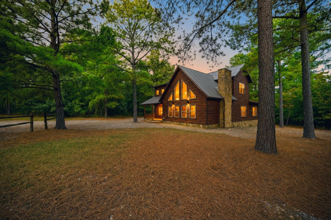A log cabin with a stone chimney, surrounded by trees and a gravel driveway.