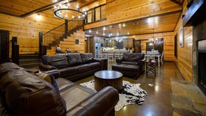 A spacious living room with a leather sofa, a round coffee table, and a fireplace. A staircase leads to an upper level, and a kitchen is visible in the background.