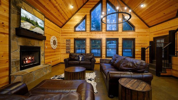 A log cabin interior with a stone fireplace, leather sofas, a round coffee table, and large windows.