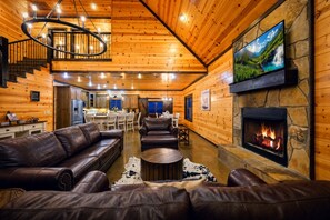 Interior de una cabaña de madera con chimenea, sofás de cuero, zona de comedor y televisor montado en la pared.