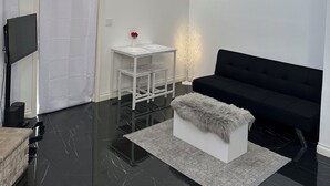 Une pièce moderne et minimaliste, avec un canapé noir, une table blanche ornée d’un vase et un téléviseur encadré de noir.