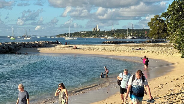 Une plage où l’on voit des gens se promener, des bateaux sur l’eau et un pont en arrière-plan.