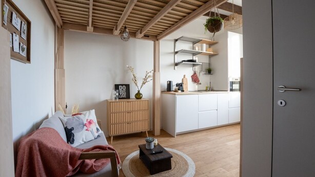 Uma sala de estar moderna com teto de madeira, um sofá com almofadas, uma pequena mesa redonda e uma área de cozinha visível por uma porta aberta.
