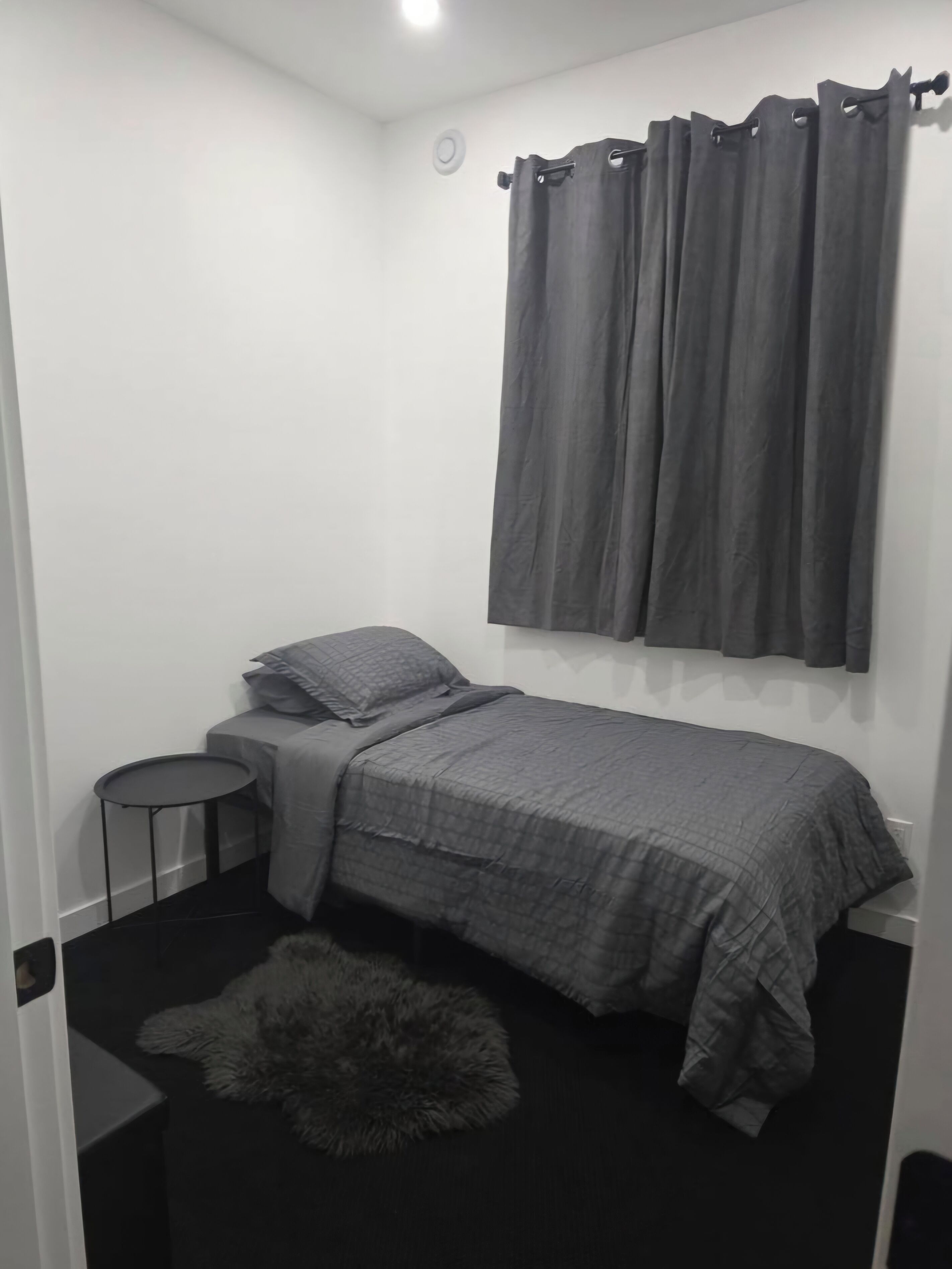 A small, dimly lit room with a single bed, a small round table, and a fluffy rug.