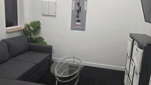 Ett modernt rum med en grå soffa, ett soffbord i glas, en liten krukväxt, en väggmonterad elcentral och en tv på väggen.