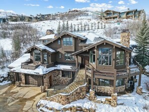 A large, multi-story wooden house with stone accents, a stone staircase, and a snow-covered landscape.