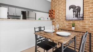 Une cuisine moderne avec des meubles blancs, une table à manger en bois mise avec des assiettes et des verres, et un mur en briques orné d’un tableau représentant un cheval.