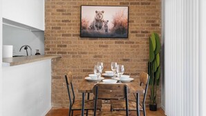 Un comedor con pared de ladrillo, un cuadro enmarcado de leones, una mesa de madera dispuesta con platos y vasos blancos, y una planta en maceta.