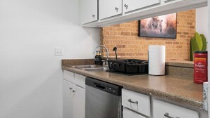 Una cocina con armarios blancos, encimera de granito, lavavajillas, fregadero con grifo, dispensador de jabón, portarrollos de papel y pared de ladrillo con un cuadro enmarcado.