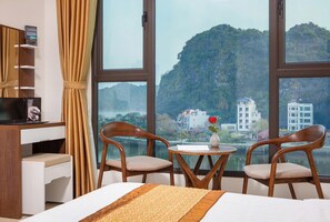 A hotel room with a bed, two wooden chairs, a small table, a TV, and a view of a mountainous landscape through large windows. - Tung Quang hotel (Hai Phong)